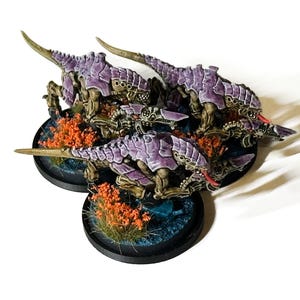 May include: Miniature figures of insectoid creatures with purple armor and sharp claws. The figures are on circular bases with orange foliage and blue accents. The figures are painted with detailed shading and highlights.