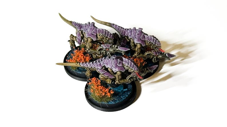 May include: Miniature figures of insectoid creatures with purple armor and sharp claws. The figures are on circular bases with orange foliage and blue accents. The figures are painted with detailed shading and highlights.