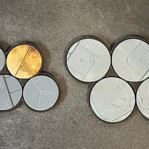 May include: A collection of round, gray miniature bases with black rims. Some bases have a cracked texture, while others have a gold, metallic finish. These bases are designed for miniature figures.