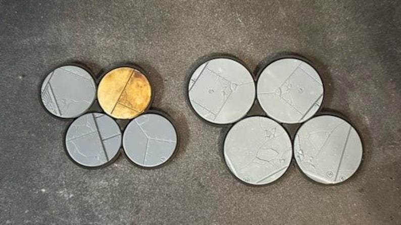 May include: A collection of round, gray miniature bases with black rims. Some bases have a cracked texture, while others have a gold, metallic finish. These bases are designed for miniature figures.