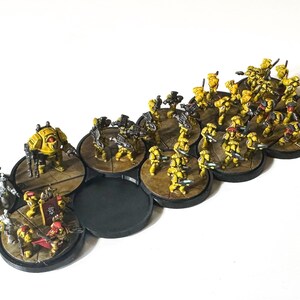 May include: Miniature figurines of a yellow and brown army, arranged on circular bases. The figures include soldiers, a tank-like vehicle, and other military units. The bases are connected, and the overall scene suggests a tabletop war game.
