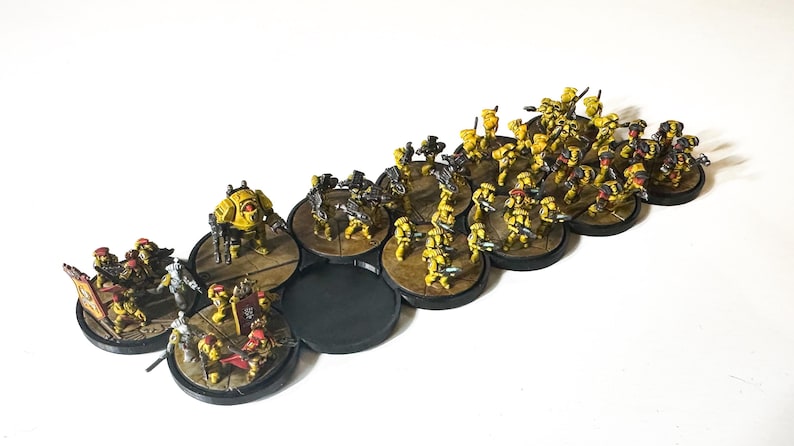 May include: Miniature figurines of a yellow and brown army, arranged on circular bases. The figures include soldiers, a tank-like vehicle, and other military units. The bases are connected, and the overall scene suggests a tabletop war game.