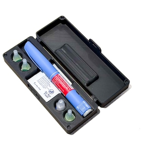 May include: A black plastic carrying case containing a blue and red medical injection pen and four small, clear, conical needle caps. A white rectangular label is visible inside the case with the text "Do Not Flush" and "1-Ply Pen".