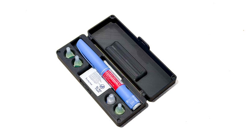 May include: A black plastic carrying case containing a blue and red medical injection pen and four small, clear, conical needle caps. A white rectangular label is visible inside the case with the text "Do Not Flush" and "1-Ply Pen".
