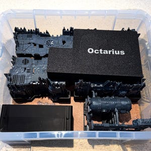 May include: A clear plastic storage bin filled with gray miniature terrain pieces and a black box labeled "Octarius." The bin contains various detailed structures, including what appears to be a tank and other industrial components.