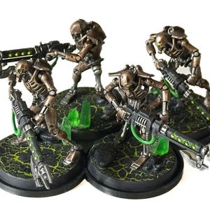 May include: Miniature figurines of Necron warriors, metallic bronze in color, with detailed skeletal structures and weaponry. The figures are posed on textured bases with green accents, set against a white background. The miniatures are designed for tabletop gaming.