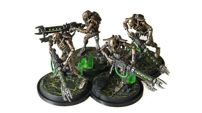 May include: Miniature figurines of Necron warriors, metallic bronze in color, with detailed skeletal structures and weaponry. The figures are posed on textured bases with green accents, set against a white background. The miniatures are designed for tabletop gaming.