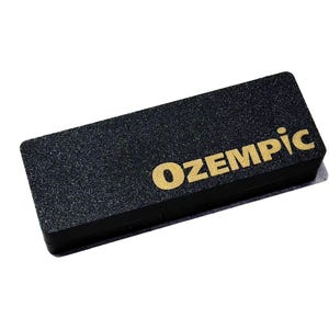 May include: A rectangular black object with a textured surface featuring the word OZEMPIC printed in gold lettering on the top right side. The item has a solid, flat construction with sharp, defined edges and a matte finish across the entire surface.