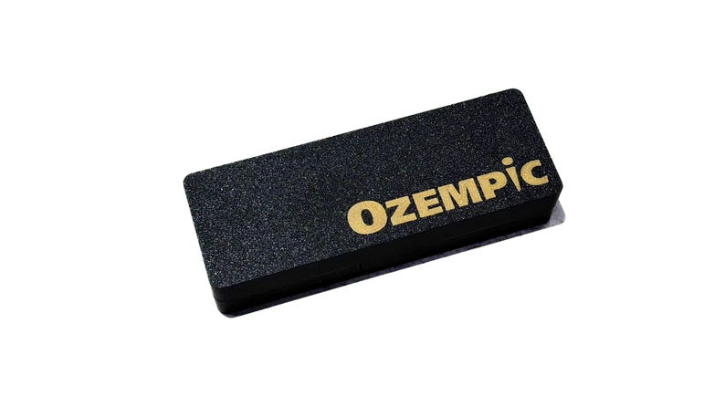 May include: A rectangular black object with a textured surface featuring the word OZEMPIC printed in gold lettering on the top right side. The item has a solid, flat construction with sharp, defined edges and a matte finish across the entire surface.