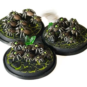 May include: Three miniature figurines of insect-like creatures with metallic brown bodies and glowing green eyes. They are clustered on circular black bases with a textured, cracked green surface, and a green crystal.