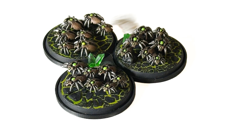 May include: Three miniature figurines of insect-like creatures with metallic brown bodies and glowing green eyes. They are clustered on circular black bases with a textured, cracked green surface, and a green crystal.