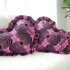 May include: Two heart-shaped decorative pillows with a pink and black geometric pattern. The pillows have a ruffled edge and are placed on a white faux fur surface. The pillows are on a white couch.
