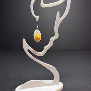 May include: A white, abstract earring display stand holding a single earring. The earring features a small, oval-shaped pendant with a yellow flower design. The stand has a curved, artistic silhouette and sits on an oval base against a dark background.