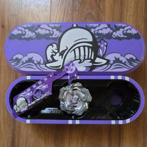 May include: Purple and black Beyblade X storage case with a white and black graphic of a knight's helmet on the lid. The case is open and shows a silver Beyblade inside.