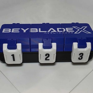 May include: A rectangular blue and black storage case with the text BEYBLADE X printed in white on the lid. The case features three white latches on the front, each labeled with a black number: 1, 2, and 3, indicating separate compartments.