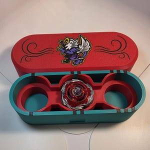 May include: A red and teal oval storage case with a lid featuring a griffin graphic and black swirl accents. The interior is teal with red inserts, holding a single red and silver circular spinning top toy in the center compartment.