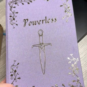 Puede incluir: Un diario de color lavanda con un diseño de lámina plateada. La portada presenta una espada decorativa y la palabra "Powerless" en una fuente de lámina plateada.