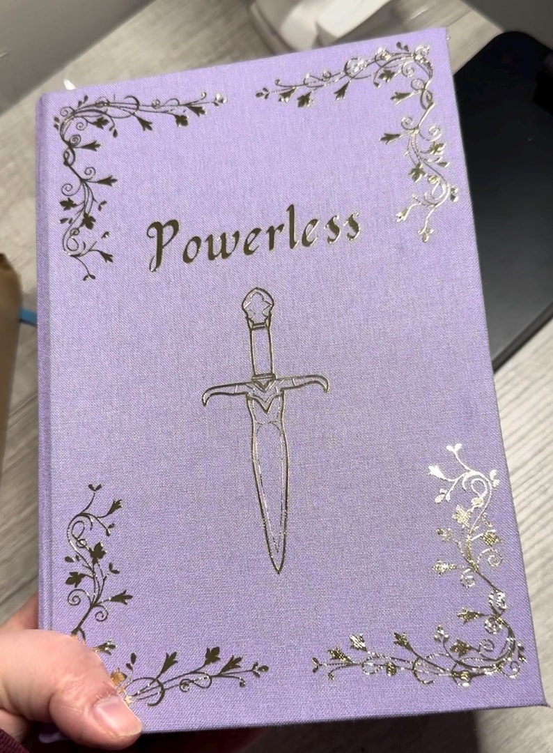 Op de afbeelding: Een lavendelkleurig dagboek met een zilveren folieontwerp. De omslag heeft een decoratief zwaard en het woord "Powerless" in een zilveren folielettertype.