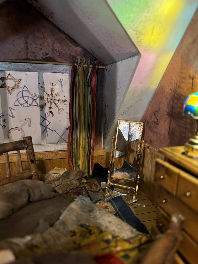 May include: A dollhouse bedroom with a bed, dresser, and a broken mirror. The walls are painted a dark brown color and have white markings. The window has a striped curtain and a view of a cloudy sky.