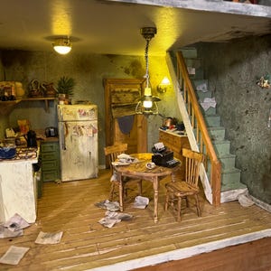 May include: A miniature dollhouse kitchen with a white refrigerator, green cabinets, and a small dining table with two chairs. The kitchen is cluttered with papers and has a worn, aged look. There is a staircase leading up to the next floor.