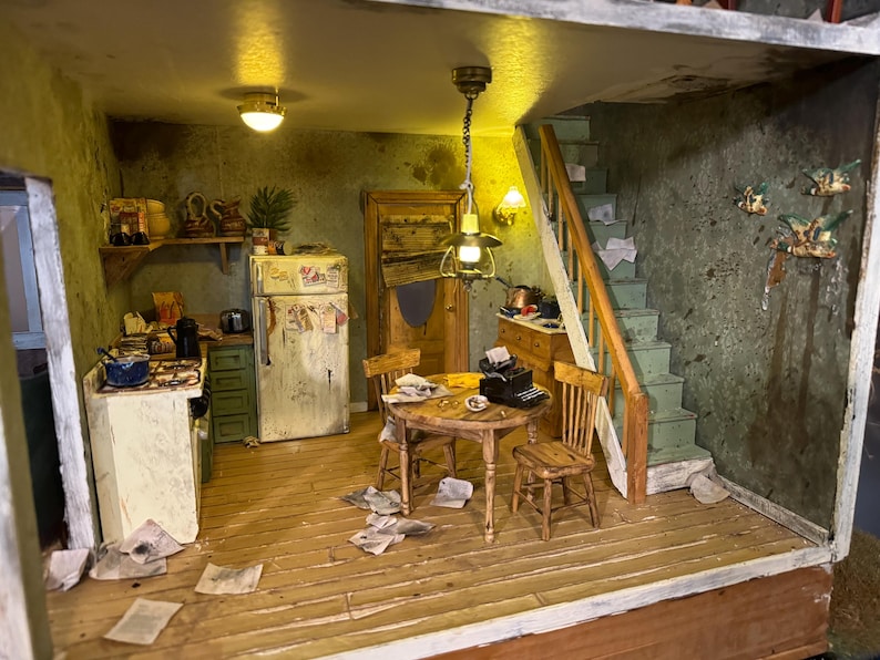 May include: A miniature dollhouse kitchen with a white refrigerator, green cabinets, and a small dining table with two chairs. The kitchen is cluttered with papers and has a worn, aged look. There is a staircase leading up to the next floor.