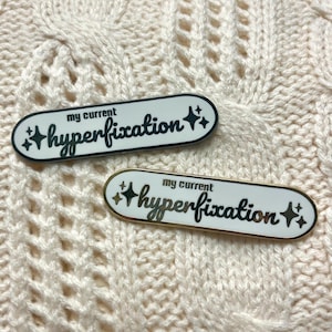 May include: Two enamel pins with a white background and black text that reads "my current hyperfixation" with gold glitter stars on either side. The top pin has a black background and the bottom pin has a gold background.
