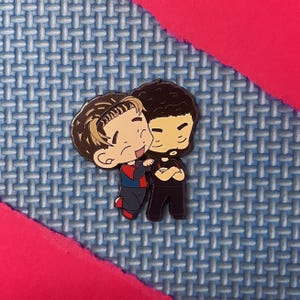Puede incluir: Un pin de esmalte metálico con dos personajes de dibujos animados en estilo chibi. Un personaje tiene el pelo castaño y viste un conjunto azul y rojo, mientras que el otro tiene el pelo oscuro rizado, barba y viste una camiseta negra.