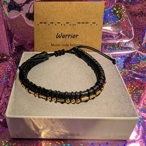 May include: Black braided bracelet with gold-colored beads and accents. The bracelet is displayed in a white box, with a card that reads "Warrior Morse code bracelet". The background is a shiny, iridescent material.
