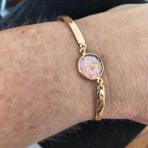 May include: Gold-toned bracelet featuring a round, iridescent charm with pink, orange, and white glitter. The bracelet has a simple, elegant design with a hinged clasp. A piece of jewelry suitable for everyday wear or special occasions.
