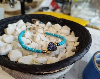 Puede incluir: Una pulsera de cuentas turquesas con una piedra triangular, morada oscura y brillante, engastada en un marco dorado. La pulsera se exhibe en una cesta negra tejida llena de piedras blancas. Cierre de nudo corredizo.