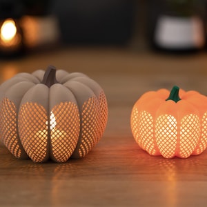 Peut inclure: Deux bougeoirs décoratifs en forme de citrouille. L'un est beige avec une tige marron, l'autre est orange avec une tige verte. Les deux ont un motif en treillis qui laisse passer la lumière, créant une lueur chaleureuse. Parfait pour la décoration d'automne.