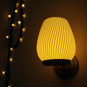 Peut inclure: Une applique murale dégageant une lumière jaune chaude. L'abat-jour en forme d'œuf est orné de nervures verticales. La base est sombre et le luminaire est monté sur une plaque circulaire de couleur bronze.