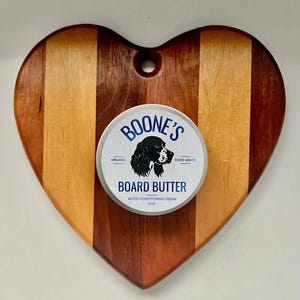 May include: Heart-shaped wooden cutting board with a striped design in shades of brown and red. A round tin container with a black and white dog illustration and the text "Boone's Organic Food Grade Board Butter Wood Conditioning Cream 6 oz" is placed on the board.