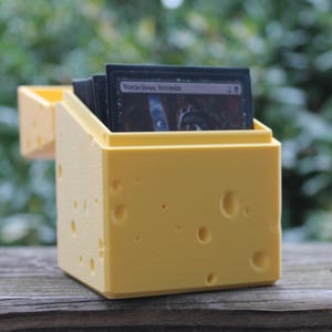Può includere: Un porta carte giallo con un design a forma di formaggio, con diversi fori rotondi. Il supporto è aperto nella parte superiore, rivelando una pila di carte nere con la scritta "Voracious Vermin". Il supporto è appoggiato su una superficie di legno.