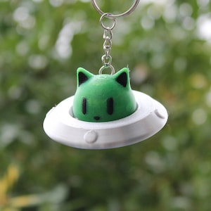 May include: A green cat-shaped figure with black ears and eyes sits inside a white UFO-shaped keychain charm. The charm is suspended from a silver keyring. The background is a blurred green.