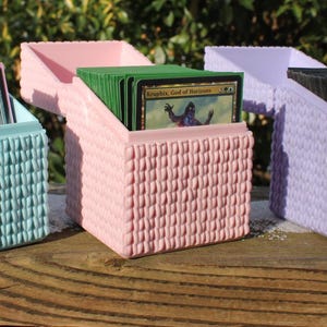 Op de afbeelding: Drie getextureerde, vierkante kaartenopbergdozen in teal, roze en lavendel, elk met een scharnierend deksel. De dozen zijn gevuld met kleurrijke ruilkaarten. De dozen worden weergegeven op een houten oppervlak.