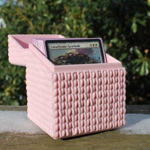 Op de afbeelding: Een roze, getextureerde kaartenopbergdoos met een scharnierend deksel, met een kaartspel. De doos heeft een geweven patroon en staat op een houten oppervlak. De bovenste kaart luidt "Cataclysmic Gearhulk".