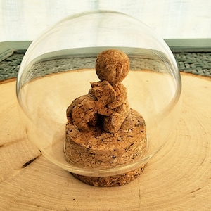 May include: A clear glass cloche with a cork base. Inside, a small cork sculpture of an elephant and a sphere sits atop the base. The cloche is on a wooden surface.
