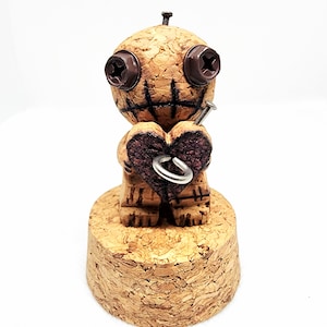 May include: A cork doll figurine with screw eyes and a nail through its head. The doll holds a heart with a metal ring. The doll is on a cork base. The doll has stitched details on its face.