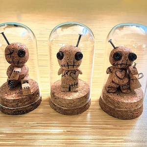 May include: Three handcrafted voodoo doll figurines inside glass cloches. The dolls are made of cork and feature button eyes, stitched mouths, and metal pins. Each doll sits on a cork base, creating a unique and artistic display.