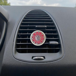 May include: A silver and red car air vent diffuser with a floral design. The round diffuser is set against a black car dashboard with air vents. The diffuser has a silver border and a red centre with a detailed silver floral pattern.