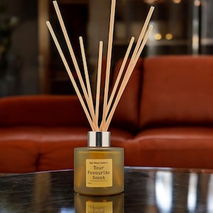 May include: A frosted glass diffuser bottle with a silver collar and wooden reeds. The bottle has a yellow label that reads "Your Favourite Scent". The diffuser is on a dark surface with a blurred red background.
