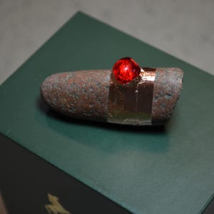 May include: A small, rough, brown stone with a red glass bead attached to it with copper tape. The stone is sitting on a dark green surface.