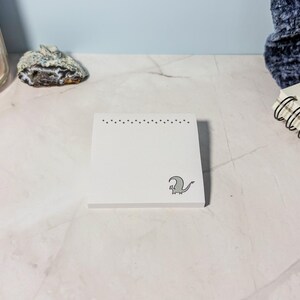 May include: A square white notepad with a gray dragon illustration in the lower right corner. The top edge features a dotted design. The notepad is on a white surface, with a candle and other stationery items in the background.