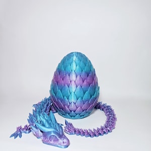 Puede incluir: Un huevo de dragón impreso en 3D con un acabado iridiscente morado y azul. El huevo está roto y un pequeño dragón con un acabado iridiscente morado y azul está emergiendo del huevo.