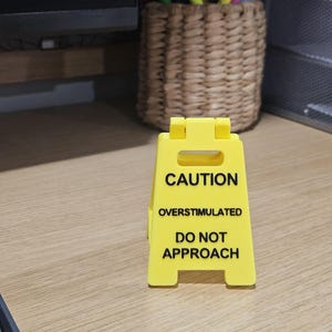 Könnte beinhalten: Ein leuchtend gelbes Warnschild mit dem Text "CAUTION OVERSTIMULATED DO NOT APPROACH" in Schwarz. Das Schild steht auf einer hellen Holzoberfläche.