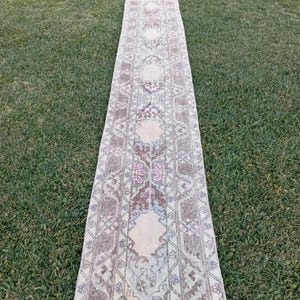 Könnte beinhalten: Ein langer, schmaler Vintage-Teppich mit verblasstem, abgenutztem Aussehen. Der Teppich weist ein sich wiederholendes Muster aus geometrischen Formen und floralen Motiven in gedämpften Tönen von Beige, Braun, Rosa und Blau auf. Der Teppich liegt auf einer grünen, grasbewachsenen Oberfläche.