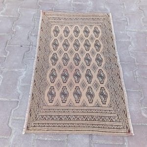 May include: Rectangular beige rug with a repeating geometric pattern in brown and grey. The rug has a detailed border and fringe along the edges. The rug is placed on a grey brick surface.