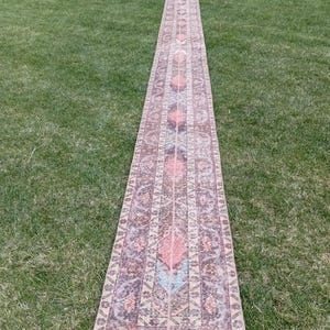 May include: A long, rectangular runner rug with a faded, vintage appearance. The rug features a central design with repeating geometric patterns in shades of pink, peach, blue, and brown. The rug is placed on a green grassy surface.