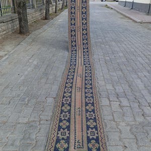 May include: A long, rectangular rug with a central peach-coloured stripe and navy blue borders. The borders feature repeating geometric patterns in white, green, and peach. The rug is displayed outdoors on a brick surface.